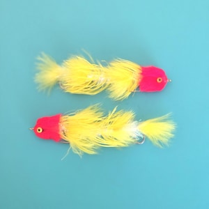 May include: Two fishing lures featuring bright pink foam heads with yellow eyes and long, fluffy yellow feathers extending along the body. Each lure is equipped with a metal hook at the end, designed for fly fishing applications.