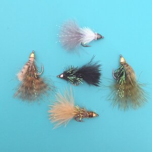 May include: Five fishing flies arranged in a cluster, featuring various colors including white, black, tan, and olive green. Each fly is constructed with soft feathers, metallic tinsel accents, and a sharp metal hook attached to a small weighted head.