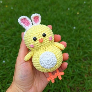 May include: A handmade yellow crochet chick with a white belly, orange feet, and a small orange beak. The chick is wearing a white headband with pink-lined bunny ears. The chick is approximately 4 inches tall.