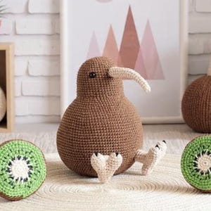 May include: A brown crocheted kiwi bird with a white beak and small feet, next to two crocheted kiwi fruit slices. The kiwi bird is on a white woven mat. A framed print with pink mountain shapes is in the background.