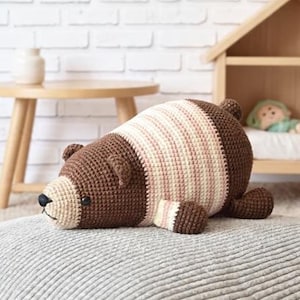 May include: A handmade brown and cream crochet bear lying on a gray textured surface. The bear has a black nose and small ears. In the background, a wooden dollhouse with a doll inside and a small wooden table with a vase.