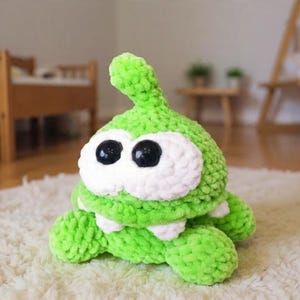 May include: A handmade green plush toy with large black eyes and white accents. The plush has a rounded body, a small mouth, and a pointed top. It is sitting on a white rug.
