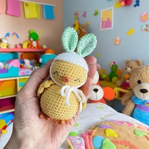 Puede incluir: Un pollito de ganchillo hecho a mano con un gorro blanco y orejas de conejo verde menta. El pollito tiene un cuerpo amarillo, patas naranjas y un lazo blanco. El juguete se sostiene en una mano, con otros juguetes visibles en el fondo.