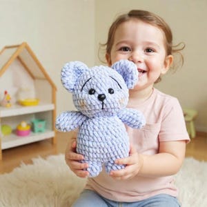 May include: A light blue crochet teddy bear with black eyes and a black nose. The bear has a smiling expression and is being held by a person. The bear is made of a soft, textured yarn.