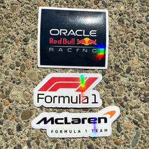 May include: Three rectangular and die-cut stickers featuring Formula 1 branding. The top sticker is black with the Oracle Red Bull Racing logo. The middle sticker is white with a red F1 logo and text. The bottom sticker is white with the McLaren logo.