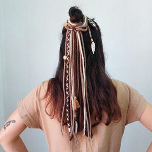 Puede incluir: Peinado femenino con moño adornado con extensiones de rastas marrones, beige y blancas. Las extensiones están decoradas con cuentas de madera, una pequeña borla y un dije en forma de hoja. La mujer lleva una camisa beige.