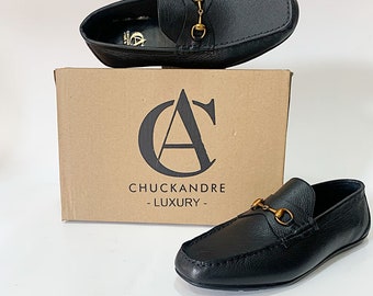 May include: A pair of black leather loafers with gold metal hardware. The loafers are on a brown box with the logo "Chuckandre - Luxury -" printed on it.