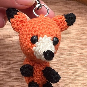 May include: A crocheted orange fox keyring with black ears, paws, and eyes. The fox has a white nose and a silver key ring.