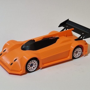 May include: An orange toy race car with a black spoiler and black windscreen. The car has white wheels and a low profile design. The toy car is a miniature replica of a race car.