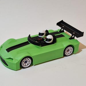 Può includere: Un'auto da corsa giocattolo verde con strisce nere e un'ala posteriore nera. L'auto ha ruote bianche e due caschi bianchi all'interno. Un giocattolo divertente per bambini.