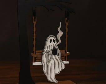 May include: A whimsical wooden decoration featuring a ghost figure sitting on a swing, holding a cup of coffee. The ghost is white with black eyes and a mouth, and the swing is suspended from a stylized tree. The design is perfect for Halloween or spooky decor.