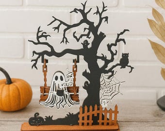 May include: A Halloween-themed wooden decoration featuring a black tree with an owl, spiderweb, and a ghost on a swing. A small pumpkin and fence are also included. The base is brown, and the background is a white brick wall.