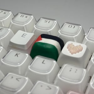 May include: Close-up of a white keyboard with custom keycaps. One keycap features the Palestinian flag, while another has a heart design. The keyboard keys are arranged in a standard layout.