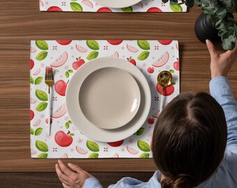 May include: A rectangular placemat featuring a repeating pattern of red apples, apple slices, and green leaves on a white background. The placemat is set with a white dinner plate, a white salad plate, a gold fork, and a gold spoon.
