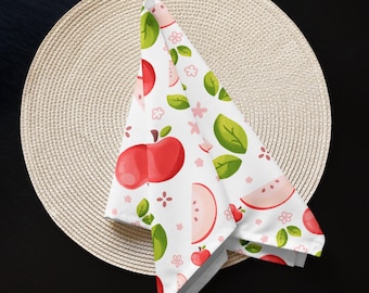 May include: A white cloth napkin featuring a repeating pattern of red apples, apple slices, green leaves, and small pink flowers. The fabric is folded into a triangular shape, displaying a vibrant fruit-themed print across the entire surface.