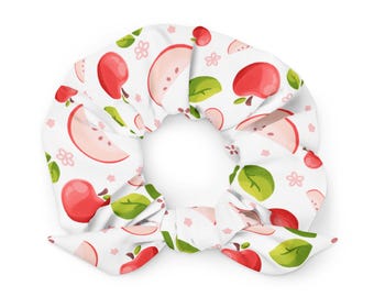 Puede incluir: Un coletero de tela blanca con un patrón repetitivo de manzanas rojas, rodajas de manzana, hojas verdes y pequeñas flores rosas. El accesorio incluye dos pequeños extremos de tela anudados en la parte inferior.