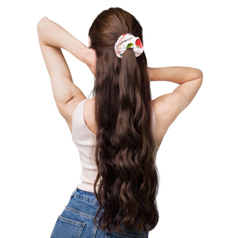 May include: A fabric hair scrunchie featuring a white background with a repeating pattern of red apples and green leaves. The scrunchie is gathered in a ruffled, circular shape and is worn in long, wavy, dark brown hair.