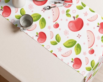 May include: A white table runner featuring a repeating pattern of red apples, apple slices, green leaves, and small pink flowers. The runner is displayed on a table with a white pitcher, two white plates, a silver spoon, and a silver fork.