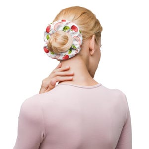 May include: A fabric hair scrunchie featuring a white background with a repeating pattern of red whole apples, sliced apple halves, and small green leaves. The scrunchie is gathered into a circular shape around a bun of blonde hair.