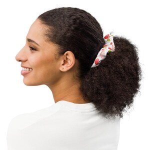May include: A white fabric hair scrunchie featuring a repeating pattern of small red apples with green leaves. The scrunchie is gathered into a circular shape and worn around a ponytail, displaying the fruit print clearly against the white background.