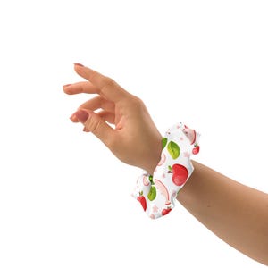 May include: A white fabric hair scrunchie featuring a repeating pattern of red apples, green apple slices, and small green leaves. The accessory has a gathered, elasticized design that creates a soft, voluminous appearance when worn.