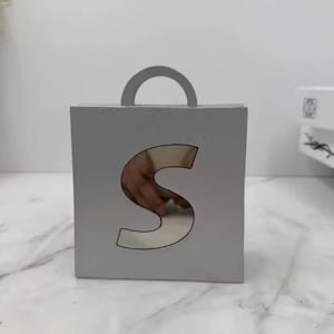 May include: A silver gift box with a handle, featuring a large cutout in the shape of the letter "S". The box is square and has a simple, modern design. The cutout reveals a glimpse of the inside.