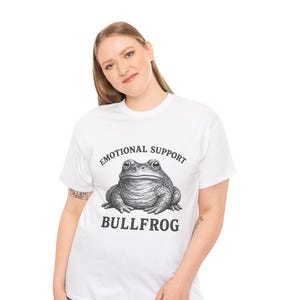 May include: A white short-sleeved t-shirt featuring a black and white graphic illustration of a bullfrog centered on the chest. Above the frog, the text reads EMOTIONAL SUPPORT, and below the frog, the text reads BULLFROG in a bold serif font.