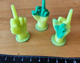 May include: Three 3D printed figurines of a middle finger gesture. The figurines are green and yellow, with the fingers painted green and the base yellow.