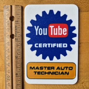 May include: A white rectangular sticker with a blue gear-shaped border. The sticker features the YouTube logo and the words "CERTIFIED" and "MASTER AUTO TECHNICIAN". A wooden ruler is next to the sticker.