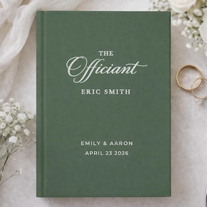 May include: A dark green wedding officiant book with the words "The Officiant" and "Eric Smith" in white script. The names "Emily & Aaron" and the date "April 23 2026" are also printed on the cover. Gold wedding rings and a white pen are visible.