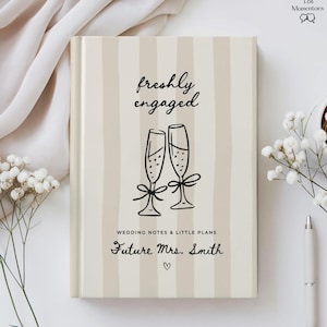 May include: A hardcover notebook with a beige and white vertical striped cover. The text reads "freshly engaged" above a line drawing of two champagne flutes. Below, it says "WEDDING NOTES & LITTLE PLANS" and "Future Mrs. Smith" with a heart.