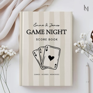 May include: A hardcover score book with a beige and white vertical striped cover. The text reads Emma & James GAME NIGHT SCORE BOOK and GAMES · SCORES · MEMORIES. A black line drawing of three playing cards with heart and diamond suits is centered.