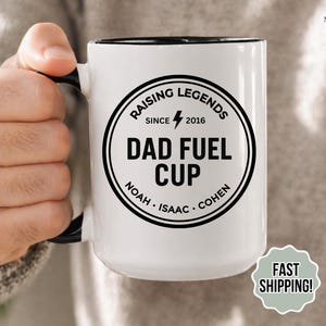 May include: White ceramic mug with a black interior and handle. The mug features a black circular design with the text "RAISING LEGENDS SINCE 2016 DAD FUEL CUP" and the names Noah, Isaac, and Cohen. A sticker reads "FAST SHIPPING!"