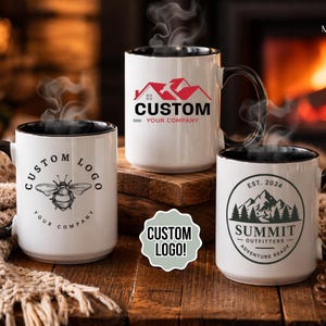 May include: Three white ceramic mugs with black handles and rims, each featuring a custom logo design. One mug displays a bee graphic, another a house logo, and the third a mountain and tree design. Steam rises from the mugs.