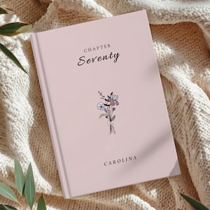 May include: A light pink hardcover journal with the words "CHAPTER Seventy" in black script. A small floral illustration is below the text. The name "CAROLINA" is printed at the bottom. The journal is resting on a cream-colored knitted blanket.