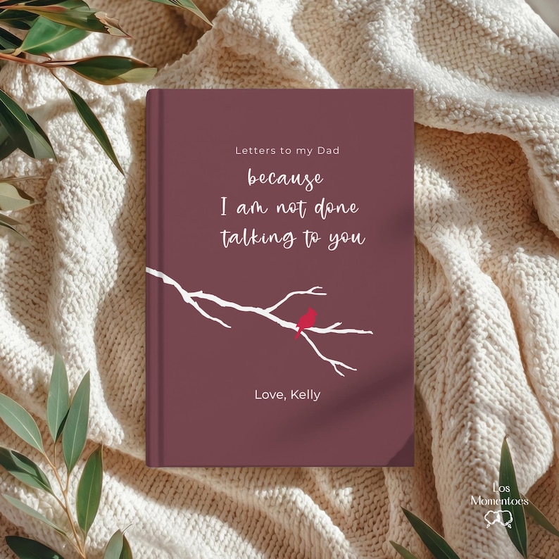 Op de afbeelding: Een bordeauxrood boek met de titel "Letters to my Dad" en de boodschap "because I am not done talking to you". De omslag toont een witte tak met een rode vogel. Het boek is gesigneerd met "Love, Kelly."