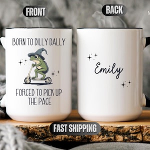 May include: White ceramic mug with a black handle. The front of the mug features a cartoon frog wearing a witch's hat and riding a scooter. The text reads "Born to dilly dally, forced to pick up the pace". The back of the mug has the name "Emily" written in cursive.