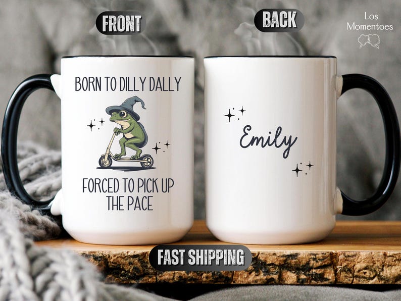 May include: White ceramic mug with a black handle. The front of the mug features a cartoon frog wearing a witch's hat and riding a scooter. The text reads "Born to dilly dally, forced to pick up the pace". The back of the mug has the name "Emily" written in cursive.