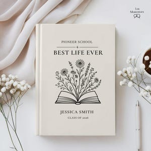 May include: A cream-colored book with the text "Pioneer School + Best Life Ever" and the name "Jessica Smith, Class of 2026." The cover features an open book with floral illustrations. A pen and a cup of coffee are on the right.