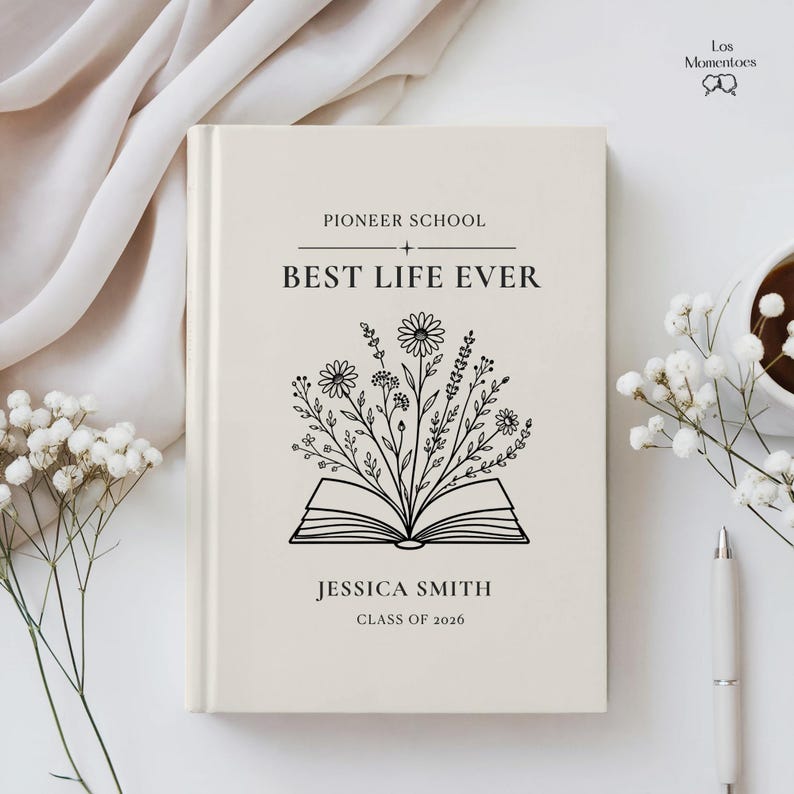 May include: A cream-colored book with the text "Pioneer School + Best Life Ever" and the name "Jessica Smith, Class of 2026." The cover features an open book with floral illustrations. A pen and a cup of coffee are on the right.