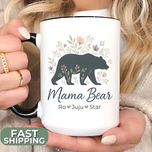 May include: White ceramic mug with a black handle and rim, featuring a dark gray bear silhouette with floral accents. The words "Mama Bear" and names are printed below the bear. The mug also has a "Fast Shipping" sticker.