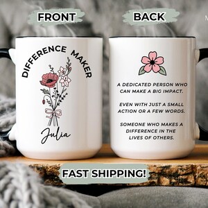 May include: White ceramic mug with a black handle and rim, featuring the text "DIFFERENCE MAKER" and a floral illustration on the front. The back of the mug has a pink flower and text. The mug is on a wooden surface.