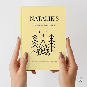 May include: A light yellow journal held by hands, with the text "NATALIE'S + CAMP MEMORIES." Below the text is a black line drawing of a campfire scene with trees and stars. The bottom of the journal reads "CAMP WILLOW LAKE &bull; SUMMER 2026."