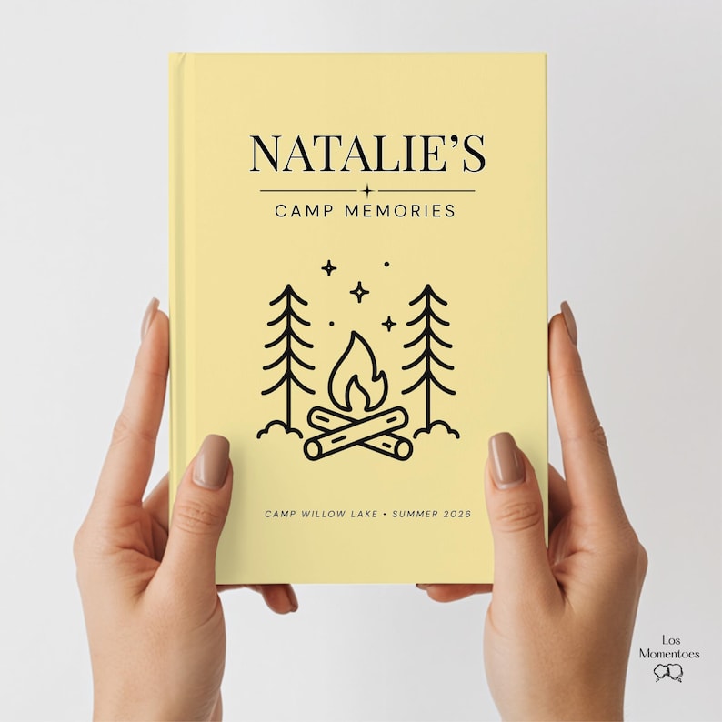 May include: A light yellow journal held by hands, with the text "NATALIE'S + CAMP MEMORIES." Below the text is a black line drawing of a campfire scene with trees and stars. The bottom of the journal reads "CAMP WILLOW LAKE &bull; SUMMER 2026."