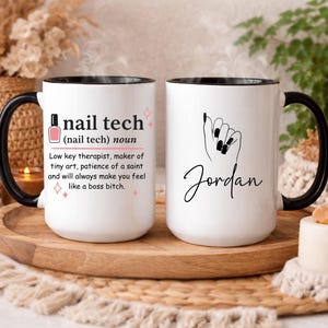 May include: Two white coffee mugs with black handles and rims. One mug features the text "nail tech" with a definition and a nail polish bottle graphic. The other mug has a hand with painted nails and the name "Jordan".