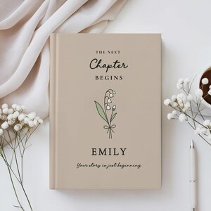 May include: A beige hardback book with the text "THE NEXT CHAPTER BEGINS" and the name "EMILY". A floral illustration is below the text. The phrase "Your story is just beginning" is at the bottom. A pen, flowers, and a cup of coffee are on the table.