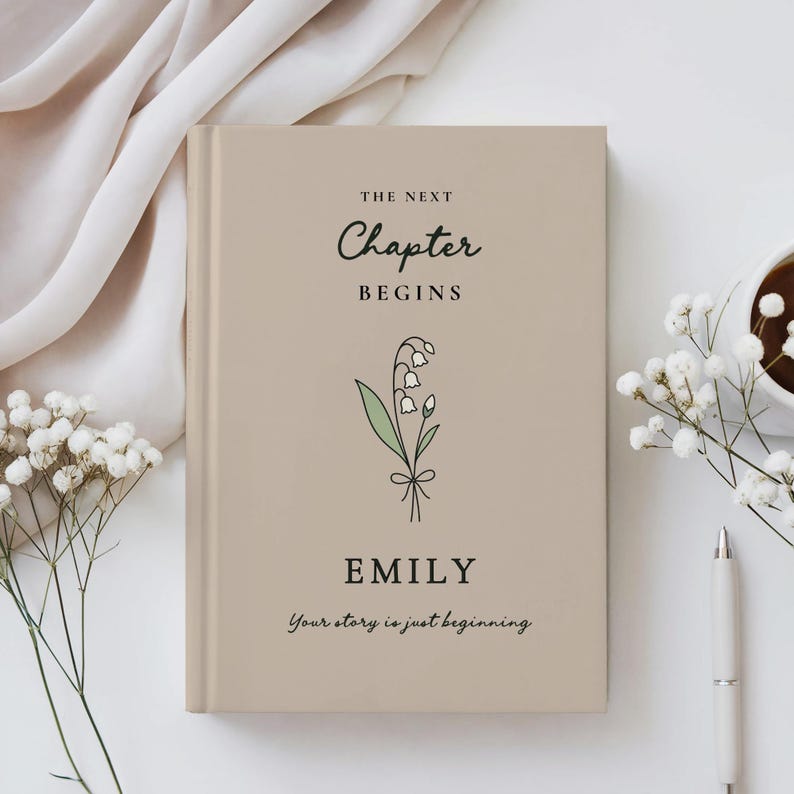 May include: A beige hardback book with the text "THE NEXT CHAPTER BEGINS" and the name "EMILY". A floral illustration is below the text. The phrase "Your story is just beginning" is at the bottom. A pen, flowers, and a cup of coffee are on the table.