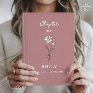 May include: A dusty rose-colored book with the word "Chapter" in cursive script at the top. Below, the word "SIXTY" is printed in capital letters. A daisy illustration is centered, and the name "EMILY" is printed below. The text "new season, beautifully written" is at the bottom.