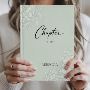 May include: A sage green journal with the word "Chapter" in elegant script. The word "Thirty" is below, and "REBECCA" is at the bottom. Floral illustrations in white adorn the corners. The journal is held by a person.