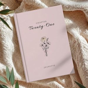May include: A light pink journal with the text "Chapter Twenty-One" in elegant script. A small floral bouquet illustration is centered below the text. The name "JEANETTE" is printed at the bottom of the journal.